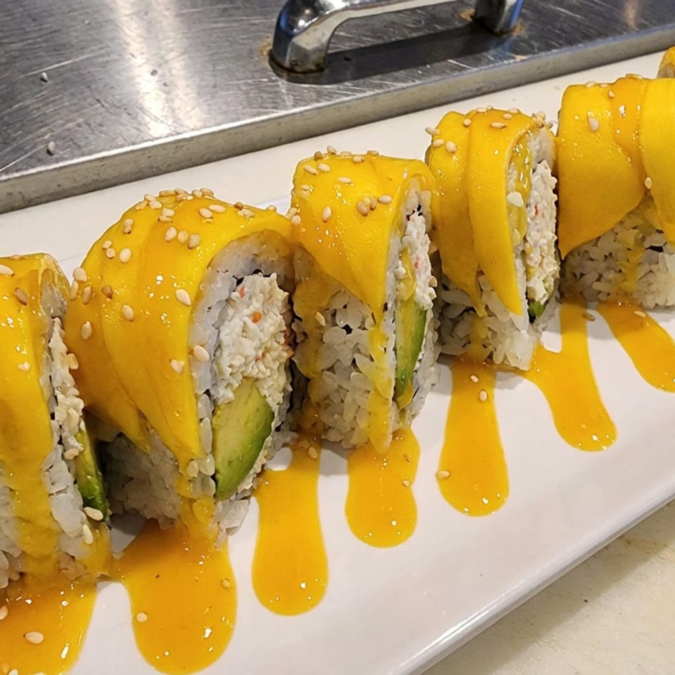 Sushi rolls topped with mango slices and sesame seeds, drizzled with golden sauce on a long white platter.
